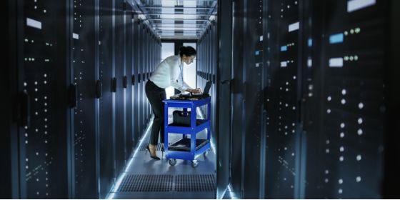 La imagen muestra a una mujer programando en un pasillo de una sala llena de servidores de datos.