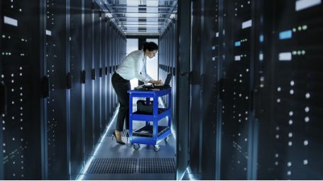 La imagen muestra a una mujer programando en un pasillo de una sala llena de servidores de datos.