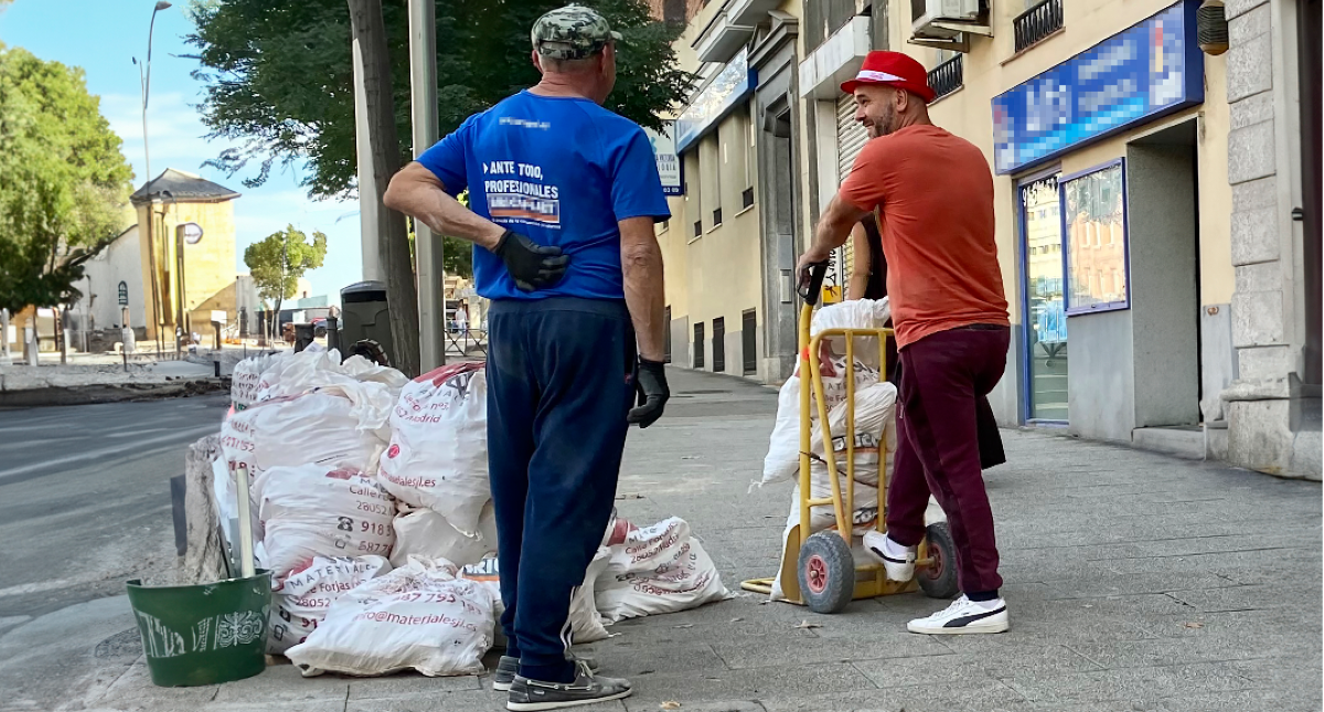 Dos trabajadores en la calle. El de la derecha lleva una camiseta naranja y un sombrero rojo con una cinta blanca y un carrito en el que transporta dos sacos. El de la izquierda lleva una camiseta azul y una gorra. Al lado de ellos se aprecia material de obra y de fondo se ven obras en la acera.