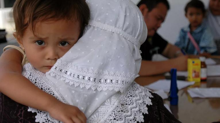 Imagen de la noticia de HIPATIA sobre permisos parentales OIT. Se ve a una niña en primer plano, cogida por su madre que está de espaldas con pañuelo en la cabeza