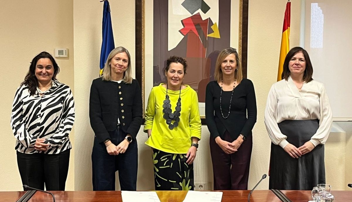 Cinco mujeres detrás de una mesa de madera en la que figuran dos carpetas, dos micrófonos y una jarra de agua. En la pared de fondo un cuadro en el centro con la bandera de Europa a la izquierda y la de España a la derecha.