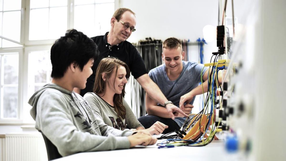 Cuatro personas mirando unos aparatos electrónicos. Tres de ellos (alumnos) sentados en una mesa y el cuarto (profesor) de pie señalando algún instrumento.